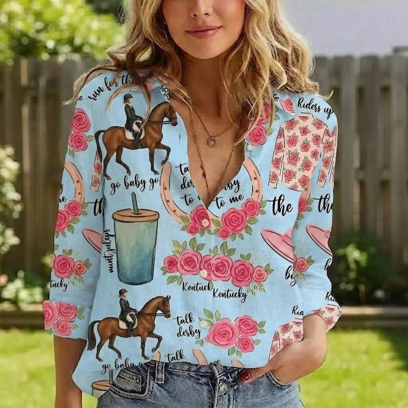 May include: A light blue long-sleeve button-down shirt featuring a repeating pattern of jockeys on brown horses, pink roses, mint julep cups, and horseshoes. The text "Riders up", "go baby go", "Kentucky", and "talk derby to me" is printed throughout.