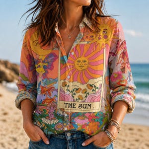 May include: A button-down shirt with a vibrant, colorful print featuring a sun, moon, and floral elements. The shirt has long sleeves and a collar. The text "THE SUN" is visible on the front. The shirt is worn on a beach.