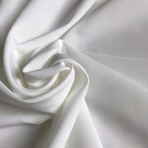 May include: A solid white fabric with a smooth, matte texture, displayed in a series of soft, flowing folds and drapes that create deep shadows and highlights across the surface, emphasizing the clean, uniform color and consistent material weave.