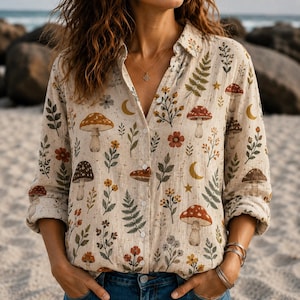 Puede incluir: Una camisa de manga larga con botones en color beige claro que presenta un patrón repetitivo de varios hongos, hojas de helecho verdes, flores pequeñas, lunas crecientes amarillas y estrellas. La camisa tiene un cuello clásico y cierre frontal.
