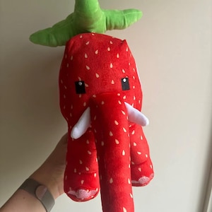 May include: A red plush elephant toy with a strawberry pattern and white seed details. It has a green leafy top, white tusks, and black square eyes. The toy is held up by a person.