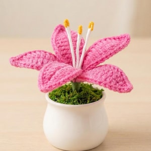 Op de afbeelding: Een handgemaakte gehaakte bloem in een kleine witte pot. De bloem heeft roze bloemblaadjes en geelgepunte meeldraden. De pot is gevuld met groen mos, wat een decoratief stuk creëert.