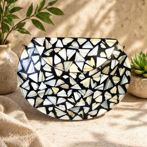 May include: A circular clutch bag with a mosaic design of black, white, cream, and yellow triangular tiles. The bag has a curved top and a thin strap. The background features a beige surface, a vase with foliage, and a succulent plant.