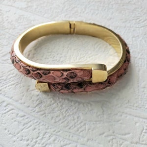 May include: A gold bangle bracelet with a pink snakeskin pattern. The bracelet is open and has a hinge on one side.