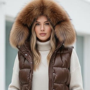Może przedstawiać: Brązowa pikowana kamizelka z kapturem obszytym futrem. Kamizelka ma pikowany wzór i zapięcie na zamek błyskawiczny. Modelka ma na sobie kremowy golf i czarne dżinsy. Futrzany kaptur jest w jasnobrązowym kolorze.