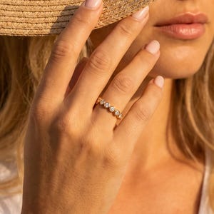 May include: A gold ring featuring a row of five small, round, clear stones set in a band. The ring is worn on a finger, with a wide-brimmed straw hat partially obscuring the face and a white top visible in the frame.