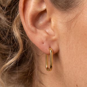 May include: A gold-toned hoop earring with a rectangular, elongated shape. The earring features a smooth, polished metallic finish and is secured through the earlobe, showcasing a clean, minimalist design with rounded edges and a continuous loop.