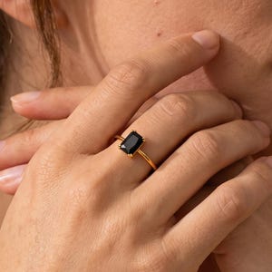 May include: A gold ring featuring a rectangular black gemstone set in a simple metal band. The stone is positioned vertically in the center of the setting, held by four small prongs, creating a clean and polished appearance on the finger.