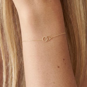 May include: Delicate gold-toned bracelet featuring two interlocking circles. The circles have a textured design, connected by a fine chain. The bracelet is worn on a wrist, showcasing its minimalist and elegant style. A simple piece of jewelry.