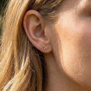 May include: A close-up of a person's ear with a gold bow-shaped stud earring. The earring is small and delicate, with a polished finish. The person has blonde hair and fair skin, with a blurred green background.