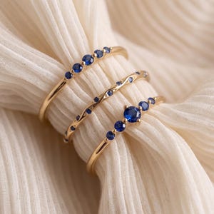 May include: Three gold rings featuring small, round, deep blue sapphire gemstones. The top ring has a row of five stones, the middle ring has a wavy band with scattered stones, and the bottom ring features a larger central stone with side accents.