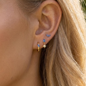 Könnte beinhalten: Nahaufnahme eines Ohrs, das mit drei verschiedenen Ohrringen geschmückt ist. Der erste ist ein kleiner Goldreifen mit einem blauen Edelstein. Der zweite ist ein goldener Ohrring mit einem Stern und einem blauen Edelstein. Der dritte ist ein kleiner Ohrring mit blauem Edelstein.
