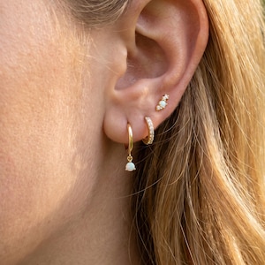 Pode incluir: Close-up de uma orelha adornada com brincos de ouro. A orelha tem três piercings, cada um com um estilo de brinco diferente: um pequeno aro com um opala pendurado, um pequeno aro com pedras transparentes e um brinco com detalhes de opala.