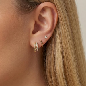 May include: A close-up of a person's ear adorned with three pieces of jewelry. The ear has a small, triangular stud earring and two gold hoop earrings, one with small, sparkling stones.