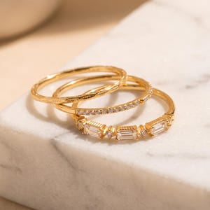 May include: A set of three gold-toned rings. One ring features a textured surface, the second is adorned with a row of small round clear stones, and the third displays three rectangular baguette-cut clear stones separated by small round stones.
