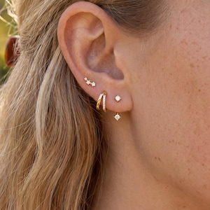 May include: Close-up of a person's ear adorned with multiple gold earrings. The ear features a double hoop, a small stud, a dangling square-shaped earring, and a cluster of three small star-like earrings. The earrings are set against the backdrop of blonde hair.