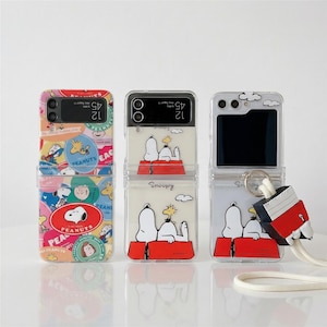 以下が含まれることがあります： スヌーピーとピーナッツのデザインが施された透明な携帯電話ケース3つ。1つのケースにはカラフルなステッカーが、他のケースには赤い犬小屋にいるスヌーピーが描かれています。白いストラップが付いたお揃いのスヌーピーチャームが1つのケースに付いています。