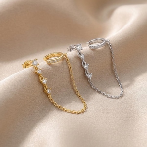 May include: Two ear cuffs with attached chains, one in gold tone and one in silver tone. Each piece features a small hoop cuff connected by a delicate chain to a stud earring, with both the cuff and the stud adorned with small, clear, sparkling stones.
