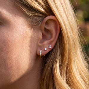 May include: Close-up of an ear displaying three different earring styles. A small gold hoop earring with clear stones, a single round stud, and a cluster of three small studs.