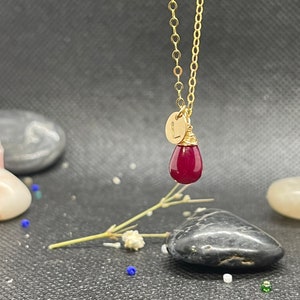 May include: A gold chain necklace with a small gold disc charm with the letter "L" engraved on it. The charm is attached to a teardrop-shaped red gemstone pendant.
