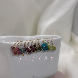 May include: A collection of six pairs of gold-colored hoop earrings, each adorned with a cluster of colorful gemstones. The gemstones vary in color, including pink, blue, purple, and green. The earrings are displayed in a white bowl, with numbers 1 through 6 below them.