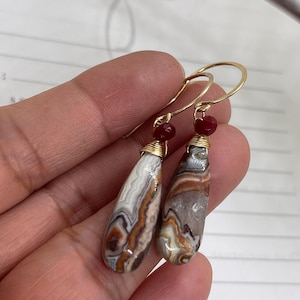 May include: A pair of teardrop-shaped earrings featuring banded agate stones with swirling patterns of brown, white, and gray. Each stone is wrapped in gold-toned wire and topped with a small, round, translucent red bead on a gold hook.