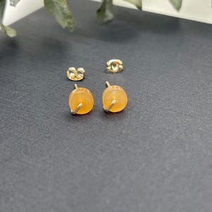 May include: A pair of acorn-shaped stud earrings with gold-colored metal accents. The earrings feature a warm, amber-colored stone and are displayed with two matching earring backs on a dark surface.