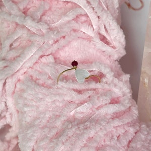 May include: A gold ring with a white heart charm and a red gemstone accent. The ring is on a pink fuzzy surface.
