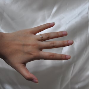 May include: A delicate gold ring with a single pearl accent. The ring is worn on the middle finger of a hand.