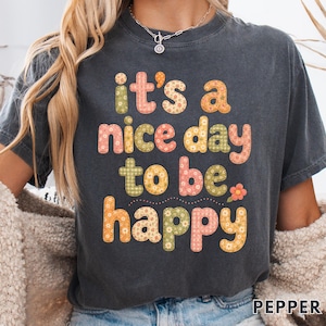 Puede incluir: Una camiseta de manga corta gris carbón con el texto "it's a nice day to be happy" en una fuente de burbujas multicolor con patrones florales. La camiseta incluye el texto "COMFORT COLORS 1717" y "PEPPER" impreso en las esquinas.