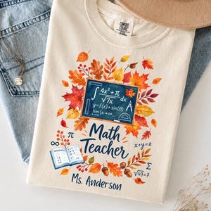Może przedstawiać: Kremowy t-shirt z grafiką tablicy z równaniami matematycznymi, otoczony jesiennymi liśćmi i żołędziami. Napis głosi Math Teacher oraz Ms. Anderson, a całość uzupełnia mała otwarta książka do matematyki i różne symbole matematyczne.