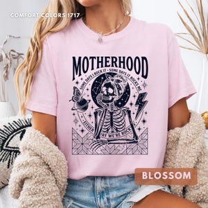 May include: A light pink t-shirt with the word "MOTHERHOOD" in black, featuring a skeleton graphic with a bandana, a butterfly, and lightning bolts. The text "SOME DAYS I ROCK IT - SOME DAYS IT ROCKS ME" is also included. The shirt is labeled "BLOSSOM".