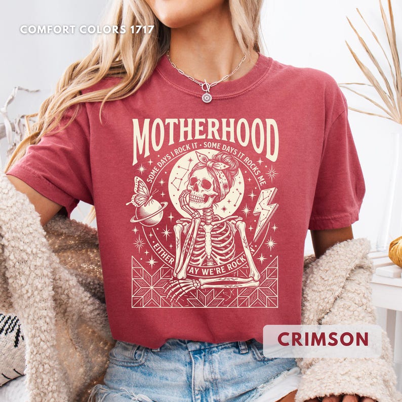 May include: Crimson-colored t-shirt with the word "Motherhood" in a vintage font. The graphic features a skeleton with a headscarf, a butterfly, and the text "Some days I rock it - some days it rocks me. Either way we're rock".
