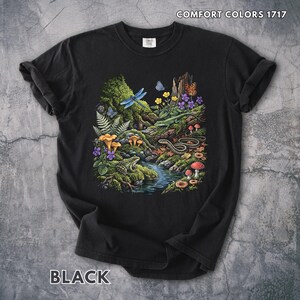 Puede incluir: Una camiseta negra con un gráfico detallado de una escena forestal con un arroyo, una rana, una serpiente, un lagarto, una libélula, mariposas, helechos, setas y flores moradas. El texto COMFORT COLORS 1717 está impreso en la esquina superior derecha.
