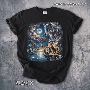 Op de afbeelding: Zwart T-shirt met een kleurrijke afbeelding van een wolf en een kleiner hondachtig wezen dat naar een volle maan kijkt. Het ontwerp bevat een nachtelijke hemel, bomen en water. De tekst "COMFORT COLORS 1717" en "BLACK" zijn gedrukt.