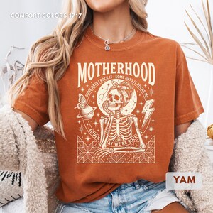 May include: A burnt orange t-shirt with the word "MOTHERHOOD" in large letters. The graphic features a skeleton wearing a bandana, a butterfly, and the text "SOME DAYS I ROCK IT - SOME DAYS IT ROCKS ME - EITHER WAY WE'RE ROCK".
