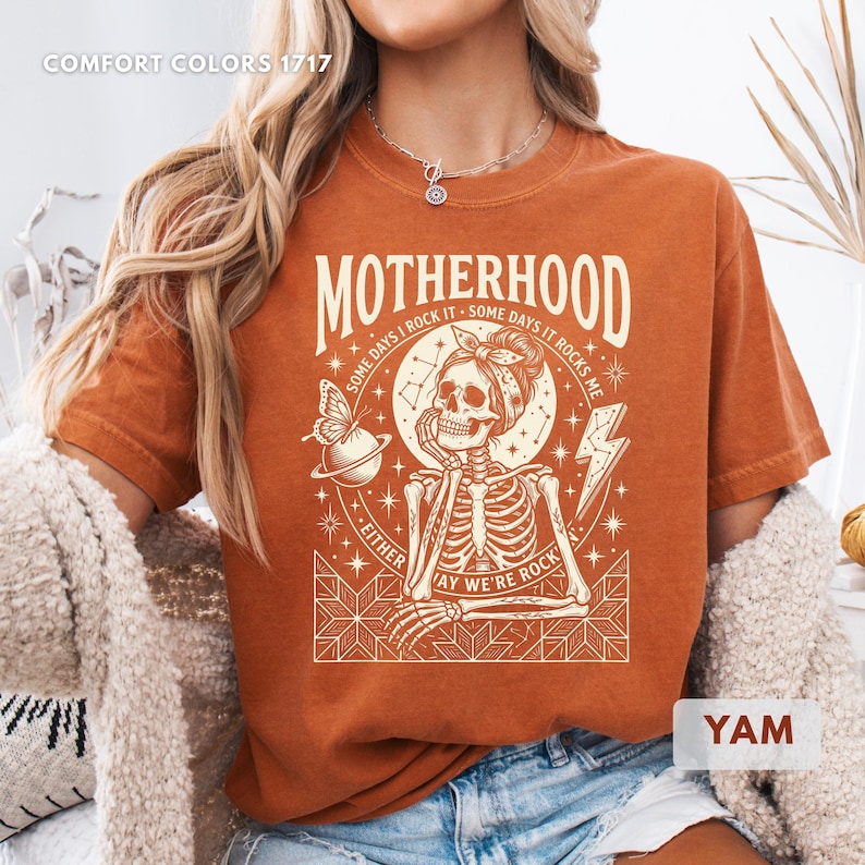 May include: A burnt orange t-shirt with the word "MOTHERHOOD" in large letters. The graphic features a skeleton wearing a bandana, a butterfly, and the text "SOME DAYS I ROCK IT - SOME DAYS IT ROCKS ME - EITHER WAY WE'RE ROCK".