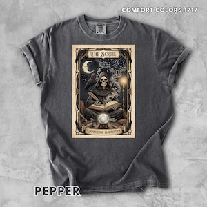 May include: A charcoal gray t-shirt featuring a graphic print of a hooded skeleton writing in a book. The design includes a crescent moon, a raven, a candle, and a crystal ball, with the text THE SCRIBE and KNOWLEDGE IS WRITTEN. The shirt is labeled COMFORT COLORS 1717 and PEPPER.