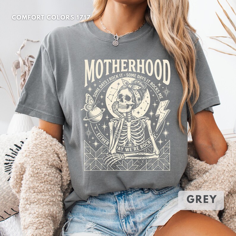 May include: Grey t-shirt with the word "MOTHERHOOD" above a skeleton graphic. The graphic includes a butterfly, a planet, a lightning bolt, and the text "SOME DAYS I ROCK IT - SOME DAYS IT ROCKS ME". The text "EITHER WAY WE'RE ROCK" is also included.