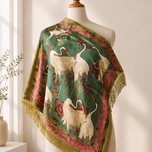 May include: A square scarf featuring a deep green background with a pattern of white cranes and red floral accents. The scarf is bordered by a red and gold decorative trim and finished with long, thin gold fringe along the edges.