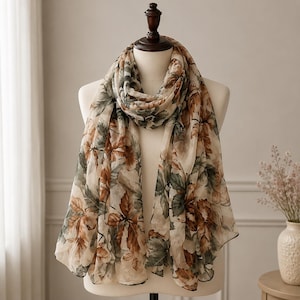 May include: A lightweight scarf featuring a botanical print with brown and muted green leaves on a cream-coloured background. The scarf is draped around a mannequin, showcasing a semi-sheer fabric with a soft, flowing texture and a repeating pattern.