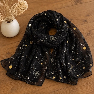 May include: A black scarf featuring a gold foil print of stars, crescent moons, and circular celestial patterns. The lightweight fabric is draped in a loose loop, showcasing the metallic gold motifs scattered across the dark, semi-sheer material.