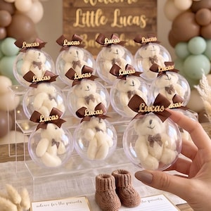 May include: Baby shower decorations with clear baubles containing teddy bears, brown bows, and the name "Lucas." A wooden sign reads "Welcome Little Lucas." The background includes balloons in gold, brown, and green, and small knitted baby booties.