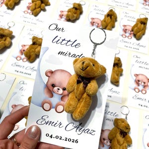 Puede incluir: Recuerdos de baby shower con pequeños llaveros de osito de peluche. Cada oso tiene un lazo marrón y está unido a un llavero plateado. Las tarjetas adjuntas dicen "Our little miracle" y "Emir Ayaz 04.02.2026".