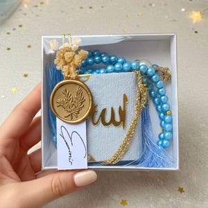 May include: A gift box containing a light blue prayer book with gold lettering, a blue beaded rosary, and a gold wax seal with a leaf design. The box is decorated with dried flowers and a small tag with handwritten script.