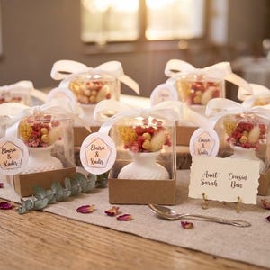 May include: Several small white textured vases inside clear boxes with brown bases, topped with white ribbons and dried pink and yellow flowers. Each box features a round tag with the text Elmira & Kadir. A place card reads Aunt Sarah Cousin Ben.