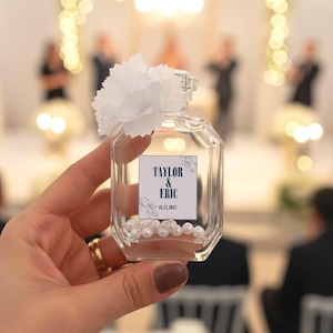 May include: A clear, octagonal glass bottle filled with white pearls, topped with a white flower, and a label that reads "TAYLOR & ERIC - 12.12.2023". The bottle is held in a person's hand, with a blurred background of a wedding.