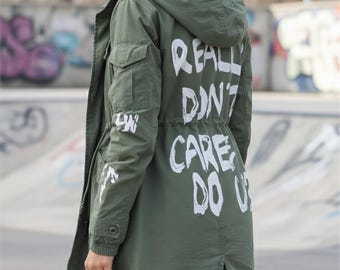 May include: An olive green hooded jacket with white text that reads "REALLY DON'T CARE DO U" on the back. The jacket has pockets and a drawstring waist. The sleeves also have white text.