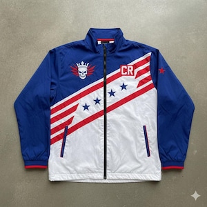 May include: A blue, white, and red zip-up jacket with a skull and crown graphic. The jacket features a diagonal stripe design with stars and the letters "CR". The jacket has a stand-up collar and two front pockets.