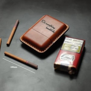 Puede incluir: Un estuche de cigarrillos de cuero marrón con el texto "Do everything beautifully" y un paquete de cigarrillos. También se ven tres cigarrillos, cada uno de unos 10 cm de largo.